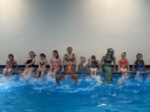 kids splashing on poolside with aquatic mermaids performers sat in between all giggling and laughing