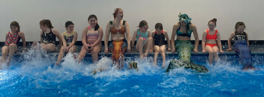 kids splashing on poolside with aquatic mermaids performers sat in between all giggling and laughi